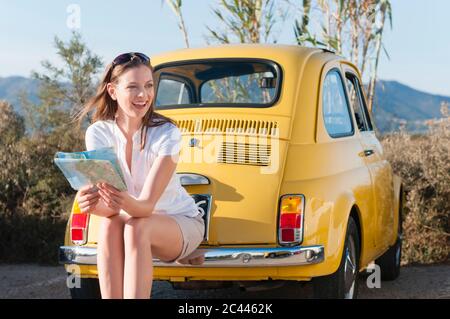 Portrait of laughing woman with map sitting on bumper of yellow vinage car, Sardinia, Italy Stock Photo