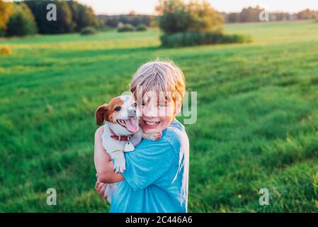 Portrait of happy boy carrying dog on field during sunny day, Poland Stock Photo