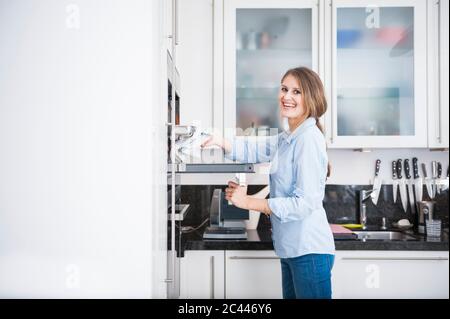 Happy beautiful young woman with long brown hair removing tray from oven Stock Photo