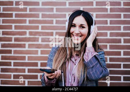 Portrait of happy woman listening music with headphones and smartphone in front of brick wall Stock Photo