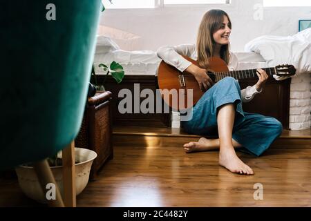 Barefoot woman sitting on the floor in front of bed playing guitar Stock Photo
