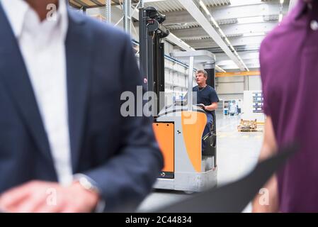 Worker in forklift in factory warehouse with two men in foreground Stock Photo