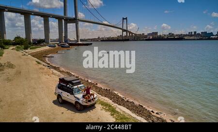 Mozambique, Katembe, Adult couple sitting on hood of 4x4 car waving at camera with city and Maputo-Katembe Bridge in background Stock Photo