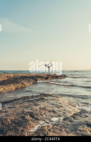 Mature woman practicing yoga at rocky beach in the evening, dancer position Stock Photo