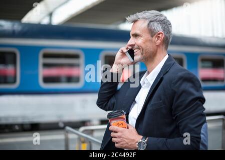 Businessman holding coffee talking over smart phone while standing at railroad station Stock Photo
