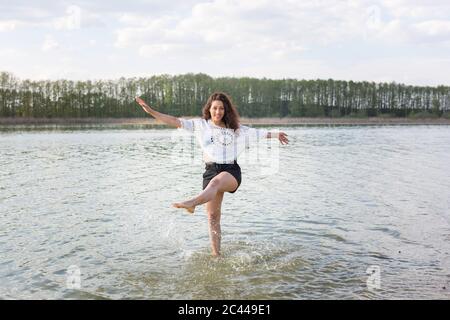 Smiling young woman standing on one leg in lake splashing with water Stock Photo