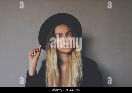 Young blond female wiccan witch wearing a black witch hat and black blazer. Artsy portrait against dark grey wall. Woman holding a hat brim Stock Photo