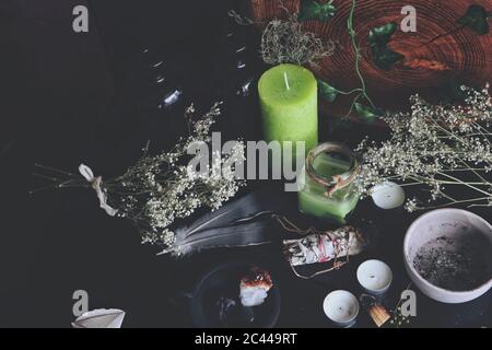 Various enchanted spell work ingredients on wiccan witch altar. Messy clutter on black table with dried baby's breath flowers, green and white candles Stock Photo
