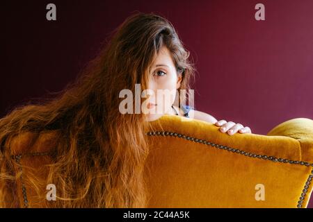 Close-up portrait of young woman with long brown hair leaning on golden chair against colored background