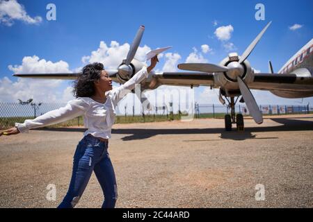 Cheerful young woman holding paper plane against propellers at airport on sunny day Stock Photo