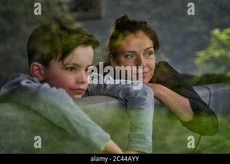 Thoughtful mother and son looking away while relaxing on sofa at home seen through window Stock Photo