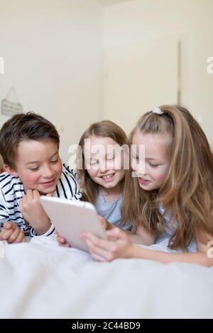Portrait of boy and his two sisters lying on bed using digital tablet for homeschooling Stock Photo