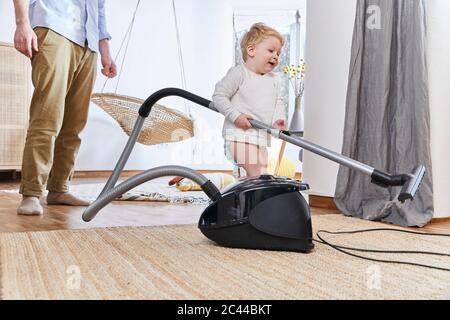 Cute baby boy holding vacuum cleaner while standing on carpet by father at home Stock Photo