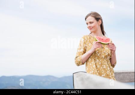 Smiling beautiful woman holding watermelon slice while standing by railing against sky Stock Photo