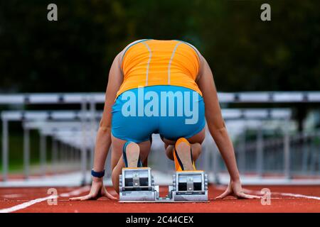 Germany, Baden Wurttemberg, Winterbach, Rear view of female hurdler kneeling at track starting block Stock Photo