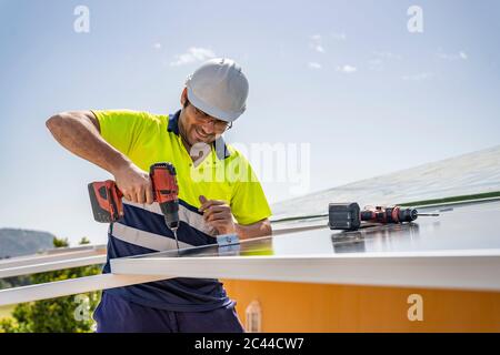 Smiling mature technician installing solar panel with drill on house roof against sky Stock Photo