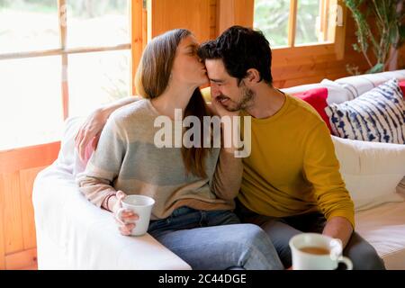 Romantic woman kissing on boyfriend's forehead while sitting over sofa in log cabin Stock Photo