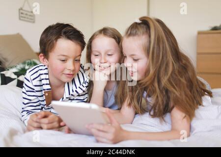 Portrait of boy and his two sisters lying on bed using digital tablet for homeschooling Stock Photo