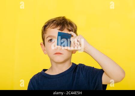Close-up of cute boy holding blue acrylic glass against yellow background Stock Photo