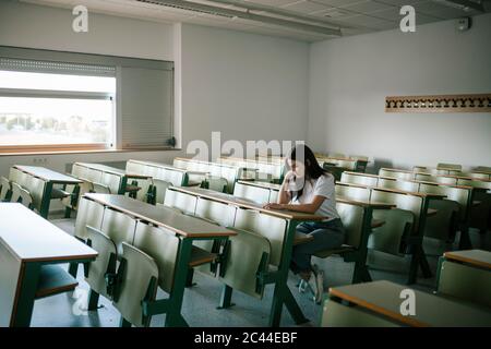 Full length of young pensive female student sitting at desk in university classroom Stock Photo
