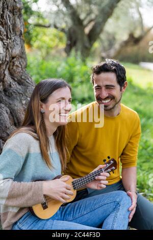 Cheerful man looking at girlfriend playing guitar while sitting in countryside Stock Photo