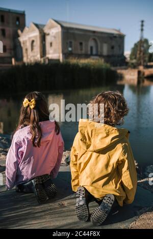 Siblings wearing raincoats while kneeling at riverbank Stock Photo