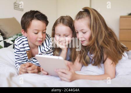 Portrait of boy and his two sisters lying on bed using digital tablet for homeschooling Stock Photo