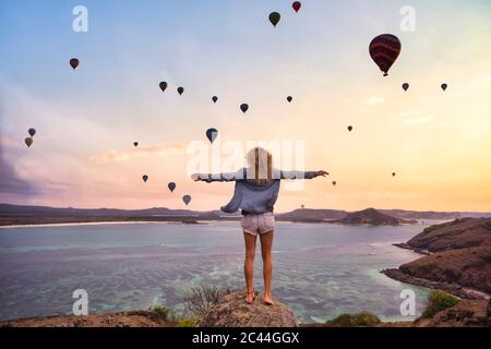 Indonesia, West Nusa Tenggara, Hot air balloons flying over lone woman standing on rocky shore with raised arms Stock Photo