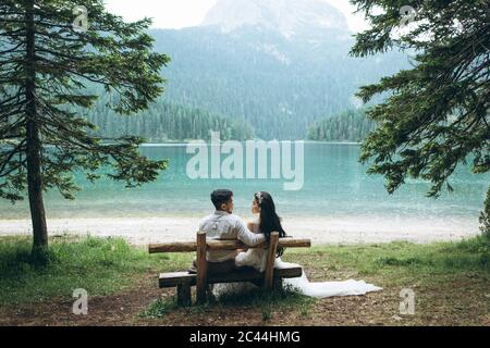 A young couple - the bride and groom are sitting on a bench on the background of the lake and looking at each other.