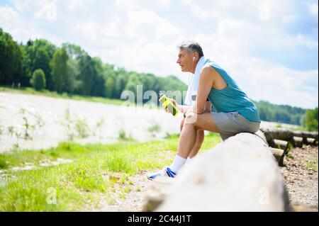 Full length side view of exhausted retired elderly man sitting on log while looking away at riverbank during sunny day Stock Photo