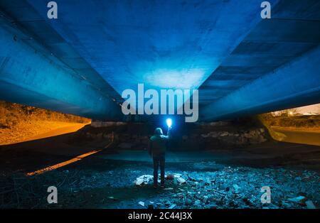 Spain, Galicia, Rear view of hooded man standing under concrete bridge with bright blue light in hand Stock Photo