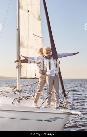 Against the wind. Happy senior couple standing on the side of sail boat or yacht deck floating in sea. Man and woman stretching hands out against the Stock Photo