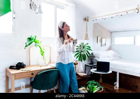 Woman standing with coffee cup while leaning on desk in bedroom at home Stock Photo