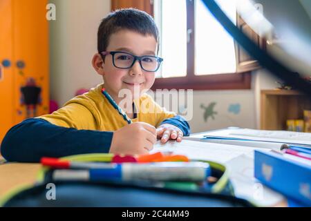 Smiling male student sitting with books at desk during coronavirus Stock Photo