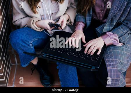 Crop view of two women sitting on stairs outdoors using laptop and smartphone
