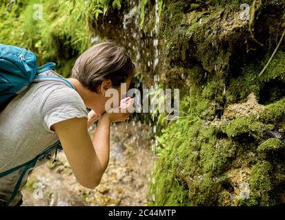 Woman drinking water from her hands cupped under trickle of Urach Waterfall in forest Stock Photo