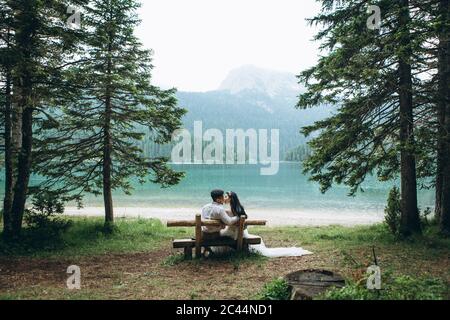A young couple - the bride and groom are sitting on a bench on the background of the lake and kissing. Stock Photo