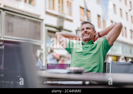 Smiling thoughtful mature man with hands behind head sitting on chair at sidewalk cafe Stock Photo