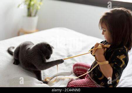 Little girl crouching on bed at home playing with cat Stock Photo