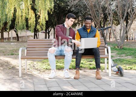 Two young men sitting on park bench sharing laptop Stock Photo