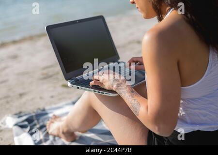 Crop view of tattooed young woman sitting on blanket on the beach using laptop Stock Photo