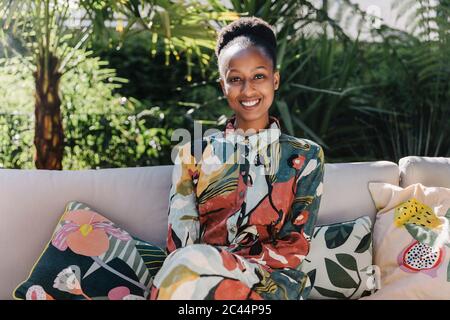 Portrait of happy young woman wearing fashionable dress relaxing on couch in garden Stock Photo