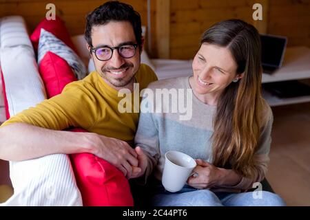 Smiling couple holding hands while relaxing on sofa in log cabin Stock Photo
