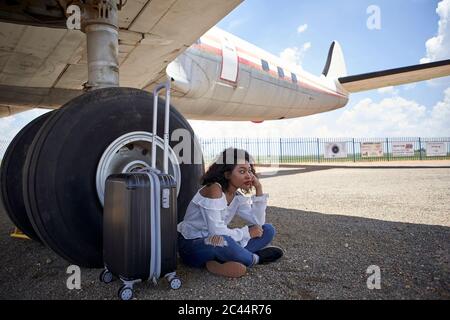 Thoughtful young woman waiting while sitting with luggage against airplane tire on runway Stock Photo