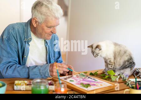 Senior man painting on paper while cat sitting on table at home Stock Photo