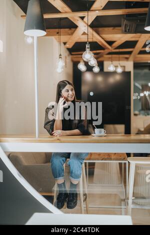 Full length of smiling beautiful young woman looking away while talking on mobile phone at cafeteria Stock Photo