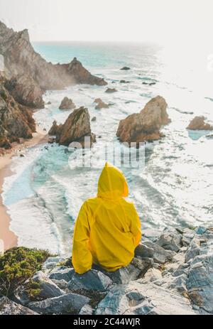 Rear view of woman wearing raincoat while sitting on rock looking at beach, Praia da Ursa, Lisboa, Portugal Stock Photo