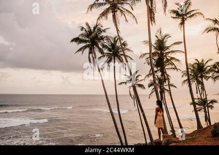 Woman looking at sea while standing by palm trees against cloudy sky, Sri Lanka Stock Photo