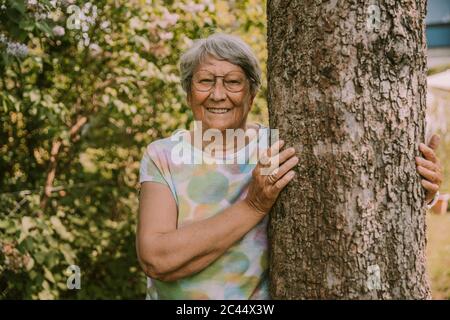 Smiling active senior woman standing by tree against plants at garden Stock Photo