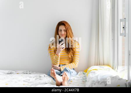 Smiling woman with long brown hair using smart phone on bed Stock Photo